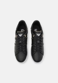 Ett par svarta lädersneakers med vita sulor, svarta snören och en vit logotyp på tungan och sidan, visade uppifrån på vit bakgrund.