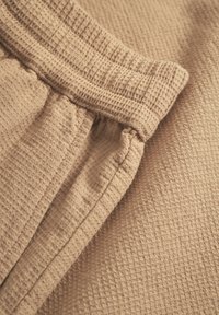 Beige waffle-textured fabric with an elastic waistband. Seam details visible at the waist and side, creating a gathered effect.