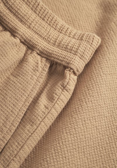 Tissu beige à texture gaufrée avec une taille élastique. Détails de couture visibles à la taille et sur le côté, créant un effet froncé.