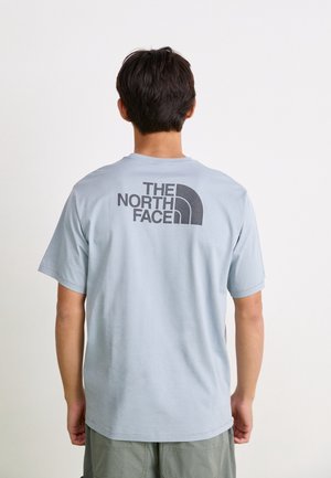 Persona di spalle, indossa una maglietta azzurra con il logo "The North Face" sul retro, in piedi davanti a uno sfondo semplice.