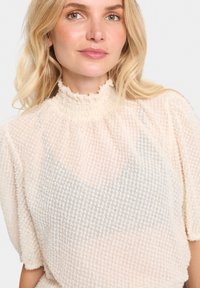 Top chiaro realizzato in tessuto texturizzato con un motivo in rilievo. Collo alto e maniche corte a sbuffo, con sottili accenti metallici.