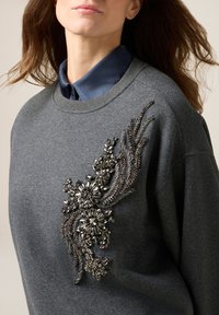 Grauer Sweatshirt mit metallischen Blumenverzierungen auf der Vorderseite. Das Design umfasst filigrane Perlenstickerei und strukturierte Akzente.