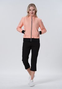 Vaaleanpunainen bomber-takki vetoketjulla, mustat yksityiskohdat hihoissa ja vyötäröllä, yhdistettynä mustiin lyhyisiin housuihin ja valkoisiin lenkkareihin.