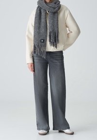 Adulto con chaqueta color crema, jeans de pierna ancha gris oscuro, zapatillas blancas y una bufanda gruesa gris con flecos.