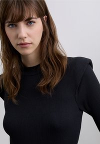 Musta pitkähihainen, resorillinen paita, jossa korkea kaulus ja jämäkät hartiat. Ominaisuuksiin kuuluu sileä pinta ja vartalonmyötäinen malli.