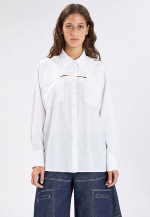 Giovane donna che indossa una camicia bianca con bottoni, con ritagli sul petto e grandi tasche frontali, abbinata a pantaloni a gamba larga in denim scuro.