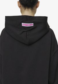 Hanorac negru cu un glugă mare și o etichetă pe spate cu branding "PGDR" roz și alb pe o patch țesută. Textură netedă de bumbac.