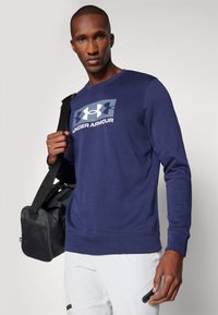 Marineblaue Sweatshirt mit Rundhalsausschnitt, Rippbündchen und großem Under Armour-Logo in Weiß und Grau auf der Vorderseite. Schwarze Sporttasche.