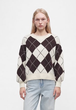 Jeune femme portant un pull crème avec un grand motif losanges noir et blanc et un jean bleu clair, debout devant un fond blanc uni.