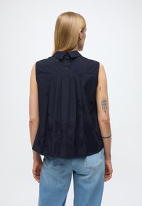 Camicia senza maniche blu navy con schiena plissettata, chiusura con bottoni al collo e texture liscia. Indossata con jeans di denim azzurro chiaro.