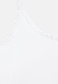 Haut blanc sans manches avec bordure en dentelle le long de l'encolure et des bretelles sur un fond uni.