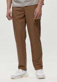 Brūnas, vieglas bikses ar gludu tekstūru, sašaurinātu dizainu un sānu kabatām, apvienotas ar baltām sporta apaviem ar pelēkiem akcentiem.