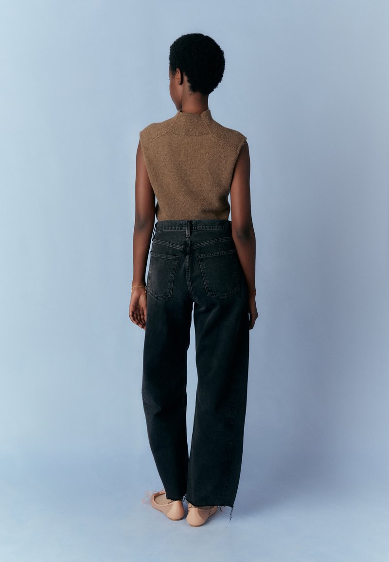 Brun ärmlös stickad topp med hög hals, matchad med högmidjade svarta raka jeans och beige ballerina skor. Enhetlig bakgrund.