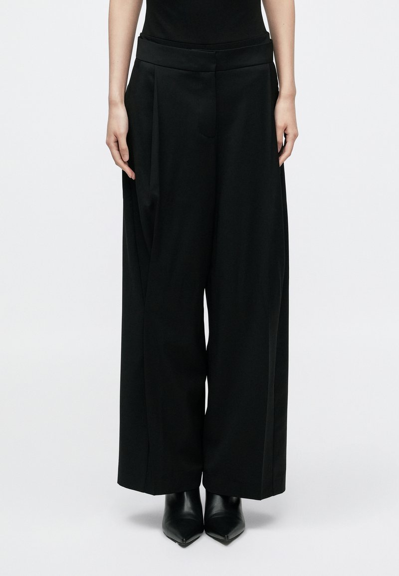 Pantalon noir à jambes larges avec plis, en tissu lisse. Comprend une ceinture plate et une poche avant, associé à des chaussures noires pointues.