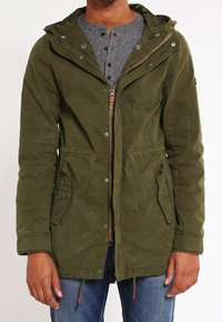 Olivgrön parka med texturerad yta, justerbar huva, dragkedja framtill och två sidofickor. Ofodrad med dragsnöre i fållen.