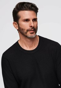 Schwarzer, strukturierter Strickpullover mit Rundhalsausschnitt und taillierter Silhouette. Das Modell hat kurzes, dunkles Haar und einen gepflegten Bart.