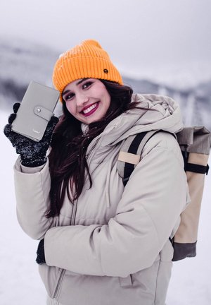Gray wallet with a button closure, held in black-gloved hand. Model wears a beige puffer jacket and orange knit beanie. Snowy background.