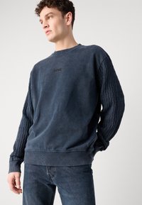 Granatowa bluza z zaciągniętymi rękawami w splot kablowy, ściągacze przy rękawach i u dołu, z subtelnym, haftowanym logo na piersi.