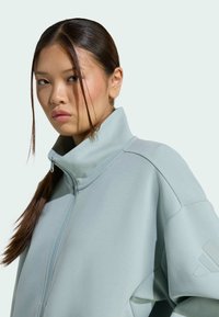 Svetlomodrá zipová bunda s vysokým golierom, s jemnou textúrou a minimalistickým dizajnom, doplnená o nenápadné logo na rukáve.