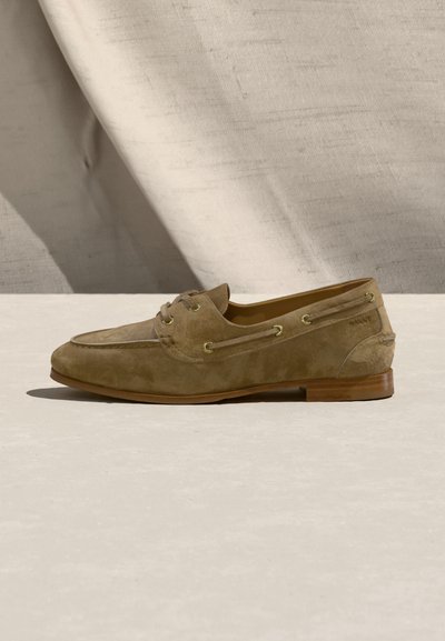 Μοκασίνι από ανοιχτό καφέ δέρμα σούεντ με κορδόνια και ξύλινη σόλα, τοποθετημένο σε ανοιχτή επιφάνεια με μπέζ υφασμάτινο φόντο.