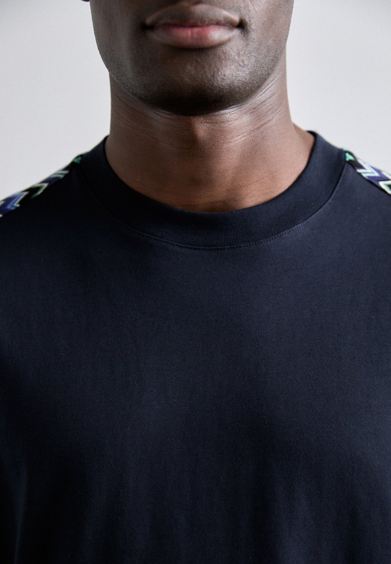 T-shirt en coton noir avec col rond côtelé et motifs en zigzag colorés sur les épaules. Texture douce avec une coupe ajustée.