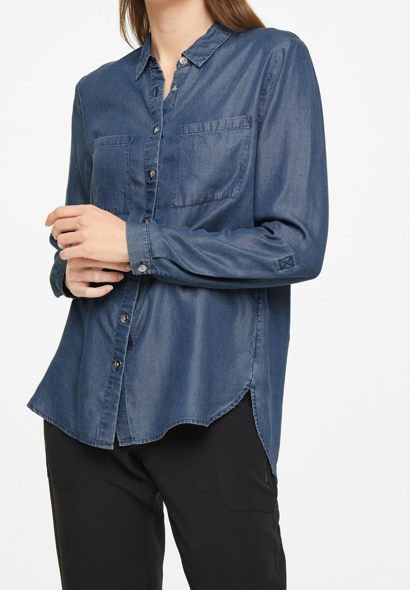 Denim shirt in dark blue, featuring a button-up design, two front pockets, and long sleeves with buttoned cuffs. Curved hem detail.