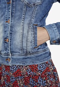 Denim jacket featuring a faded blue finish, five copper buttons, and a classic collar, paired with a floral patterned skirt.