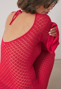 Naine kannab punast, avatud seljaga, pikade varrukatega võrkdressi, reguleerib varrukat, tal on lühikesed juuksed ja mustad prillid.