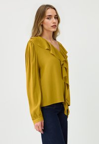 Blusa a maniche lunghe color giallo senape con dettagli arricciati sul davanti e sul collo. Tessuto leggero, polsini aderenti e siluetta morbida.