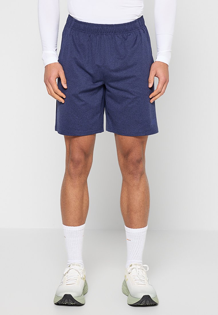 Homme portant un short bleu marine à taille élastique, un haut blanc à manches longues, des chaussettes blanches et des baskets blanches, debout devant un fond uni.