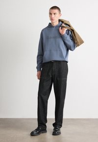 Pelēks kapuce ar "Calvin Klein" logo, brīvas tumšas denima bikses un melnas kurpes. Modelis tur brūnu jaku pār plecu.