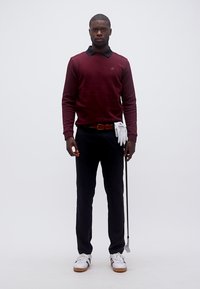 Άνδρας που στέκεται με μπαστούνι γκολφ, φοράει μπορντό πουλόβερ, μαύρο παντελόνι, λευκό γάντι γκολφ και λευκά αθλητικά παπούτσια σε απλό φόντο.