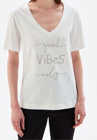 Bílá bavlněná tričko s výstřihem do V a krátkými rukávy. Má kovový nápis "dobré vibrace pouze" v hravém písmu.