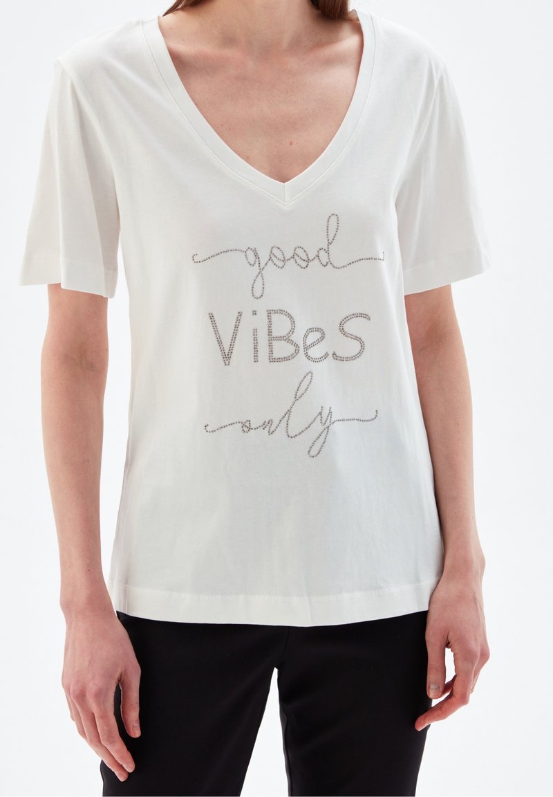 Bílá bavlněná tričko s výstřihem do V a krátkými rukávy. Má kovový nápis "dobré vibrace pouze" v hravém písmu.