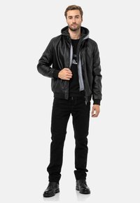 Schwarze Lederbomberjacke mit grauem Kapuzen, Reißverschluss, gerippten Bündchen und einer Fronttasche, kombiniert mit einem schwarzen Hemd und einer Hose. Schwarze Stiefel.