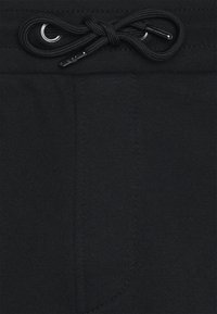 Pantalon en tissu noir avec un gros plan sur la taille, présentant un cordon noir attaché en nœud et des embouts métalliques.