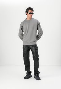 Felpa grigia in pile con spalle scese, piccolo logo rosso; pantaloni in pelle nera, strutturati, vestibilità ampia; occhiali da sole scuri e scarpe sportive nere.