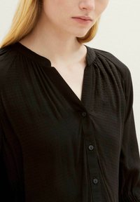 Schwarze Bluse mit platziertem Kragen und langen Ärmeln. Hat ein dezentes strukturiertes Muster und runde Knöpfe auf der Vorderseite.