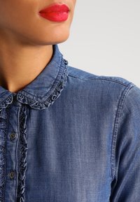 Kvinna som bär en blå jeansskjorta med volangkrage och knappslå, ljusröd läppstift och ljus hudton mot en enfärgad bakgrund.