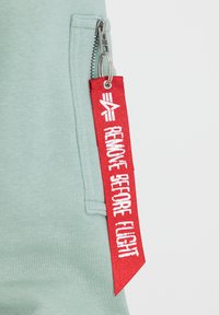 Zelena dukserica sa zatvaračem i istaknutom crvenom oznakom s natpisom "UKLONI PRIJE LETA" bijelim slovima.