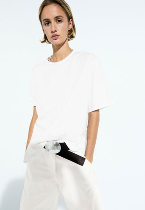 Femme aux cheveux blonds courts portant un t-shirt blanc oversize et un pantalon blanc avec une ceinture noire, debout devant un fond blanc uni.