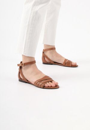 Braune Leder-Sandalen mit mehreren horizontalen Riemen, einem Knöchelriemen mit Schnalle und einer flachen Sohle. Getragen mit weißen, kürzeren Hosen.