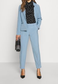 Lichtblauw op maat gemaakt pak met een getailleerde blazer en rechte broekspijpen, gecombineerd met een zwarte blouse met stippen en zwarte gepoetste hakken.