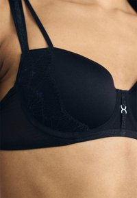 Sutiã preto com sobreposição de renda, alças ajustáveis e fecho frontal com um detalhe prateado. Tecido suave com textura detalhada.
