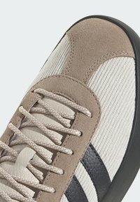 Bež i krem tenisica od brušene kože i teksturiranog tkanog materijala, ojačani vrh i ravne vezice. Na bočnoj strani nalaze se tamnosive pruge.