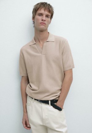 Giovane uomo che indossa una polo a maniche corte beige in maglia infilata in pantaloni bianco sporco con una cintura nera intrecciata, in piedi contro una parete chiara e liscia.