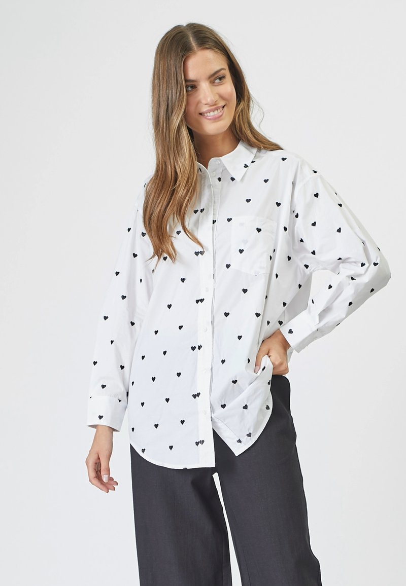 Camicia bianca con bottoni, caratterizzata da piccoli motivi a cuore neri, maniche lunghe e tasca sul petto. Indossata con pantaloni scuri.