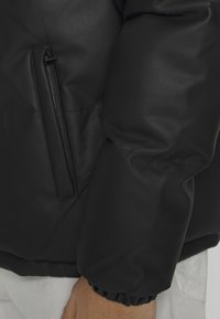 Zbliżenie na rękaw czarnej pikowanej kurtki z elastycznym mankietem i pionową kieszenią na zamek, noszonej na białych spodniach.