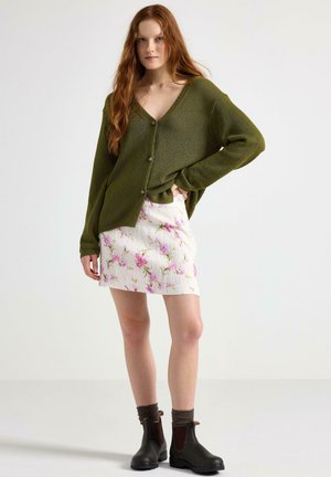 Cardigan a maglia verde oliva con scollatura a V e chiusura con bottoni, abbinato a una mini gonna bianca a fiori. Stivali alla caviglia marroni e calze grigie.