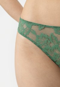 String vert en dentelle avec motif floral et maille semi-transparente. Texture lisse et douce avec une large ceinture feuillue et un bord délicat.